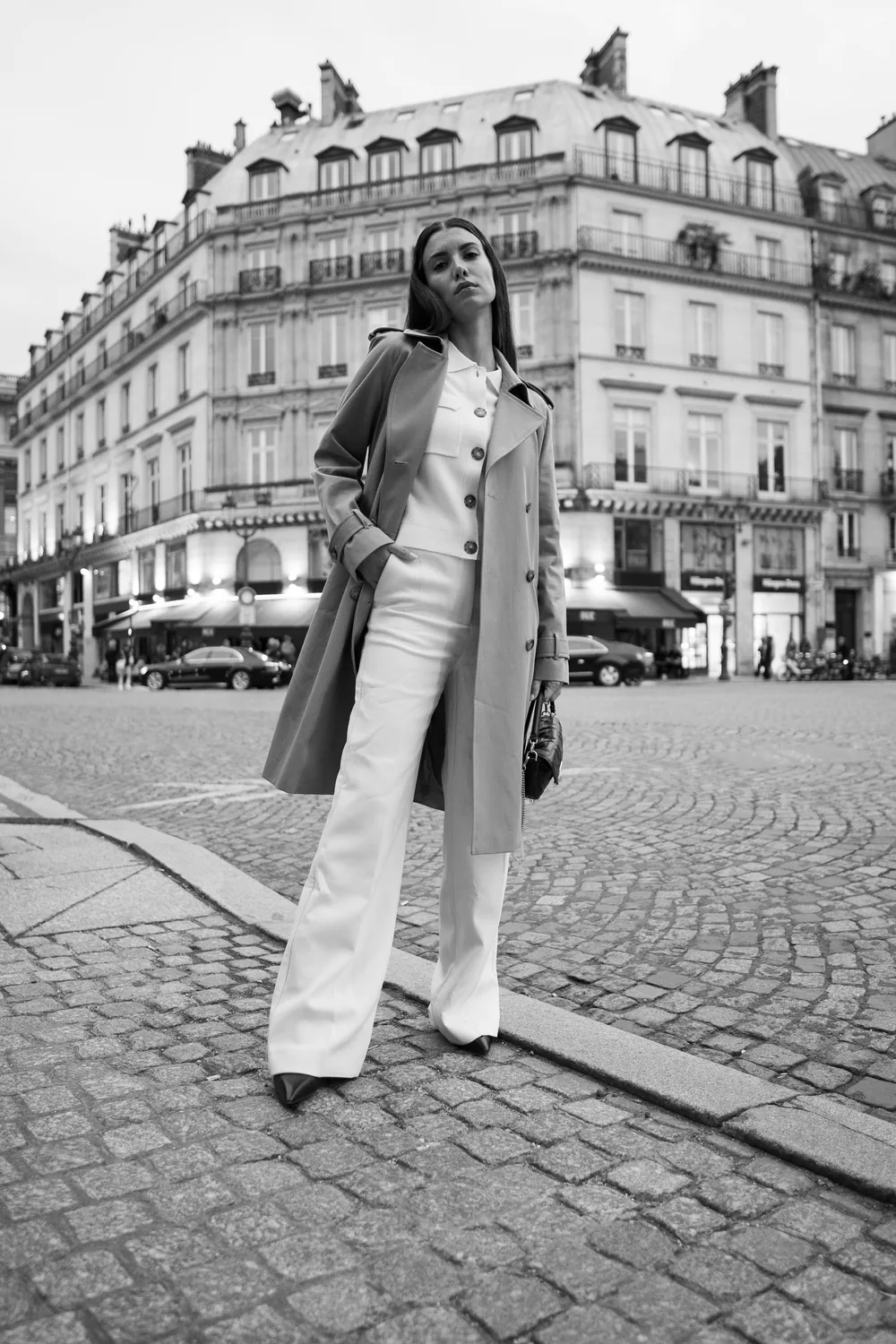 Femme en long manteau gris et pantalon blanc large sur une place pavée devant un immeuble haussmannien, en noir et blanc